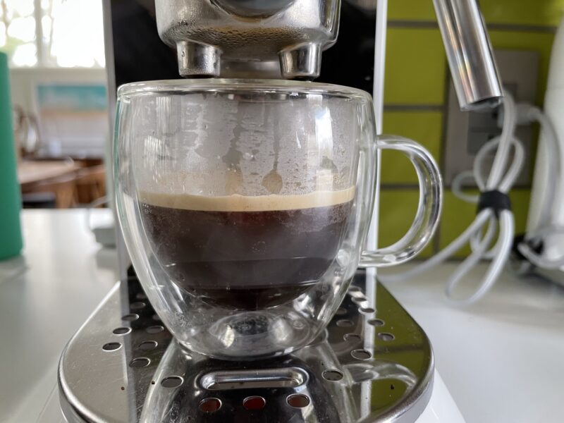 Clear espresso mug half filled with espresso, sitting on the tray of a small espresso machine